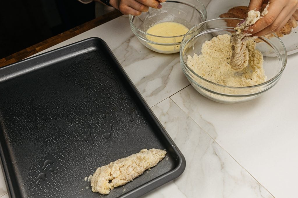 keto chicken tenders dipping chicken into the breading mixture and placing onto a baking sheet