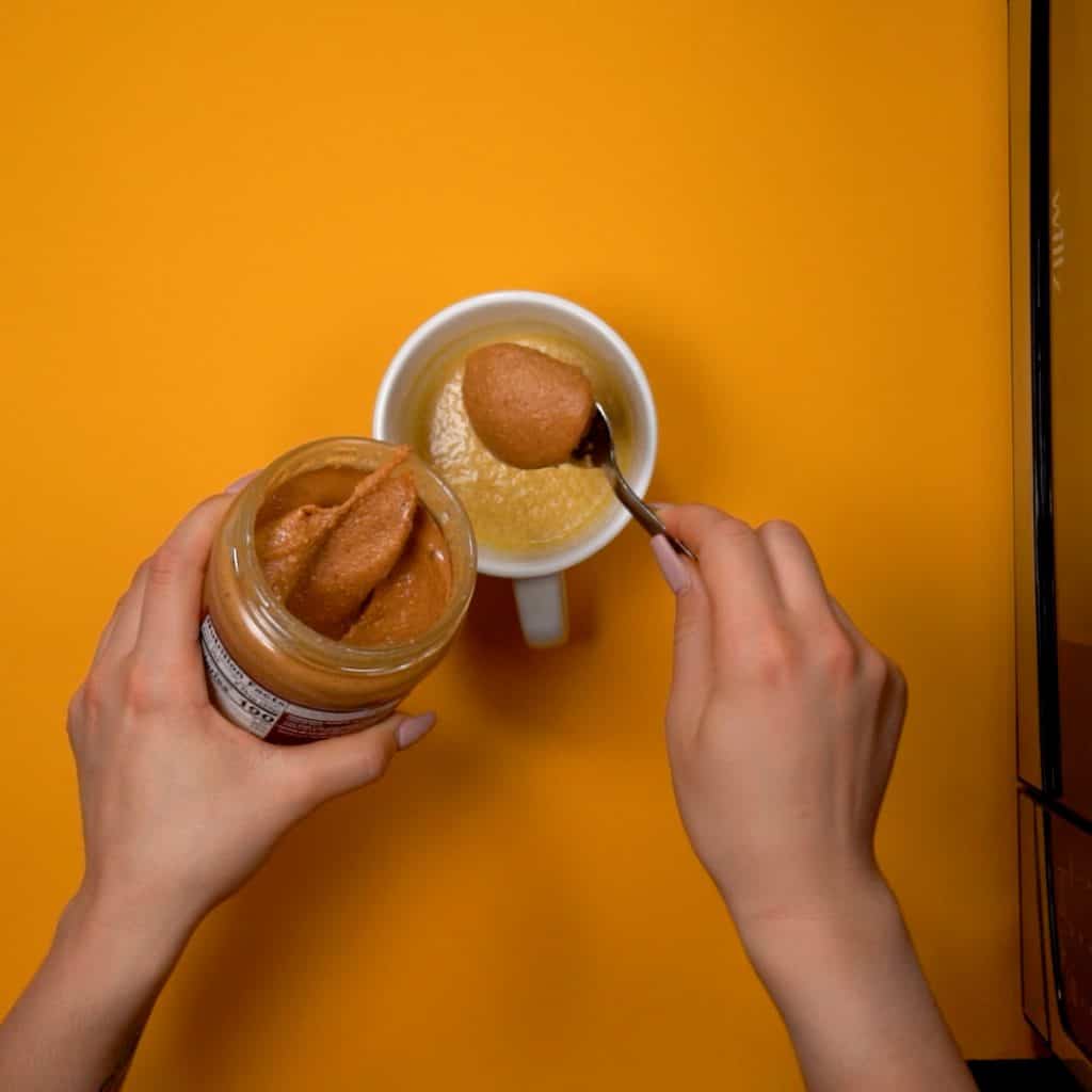 keto Mug Cake peanut butter being adding to a white batter in a coffee mug