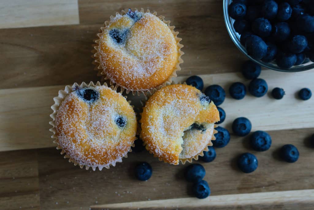 Three sweetener topped blueberry muffins on a cutting board next to blueberries