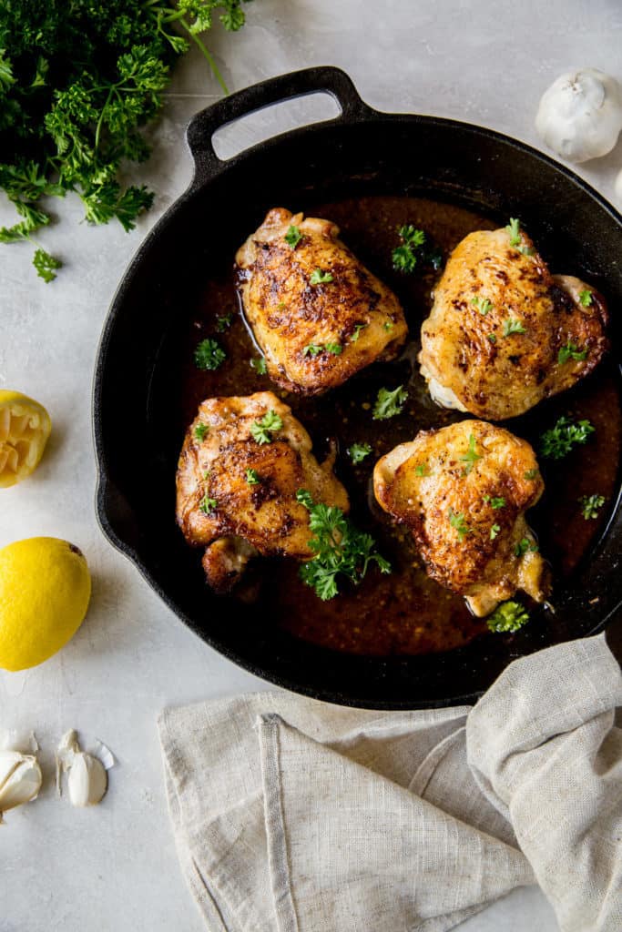 A cast iron with chicken next to lemons and garlic