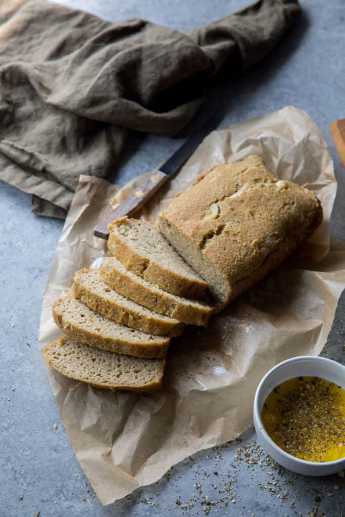 A half sliced loaf of keto garlic bread on parchment paper with a small bowl of olive oil and spices, a kinfe and dish towel