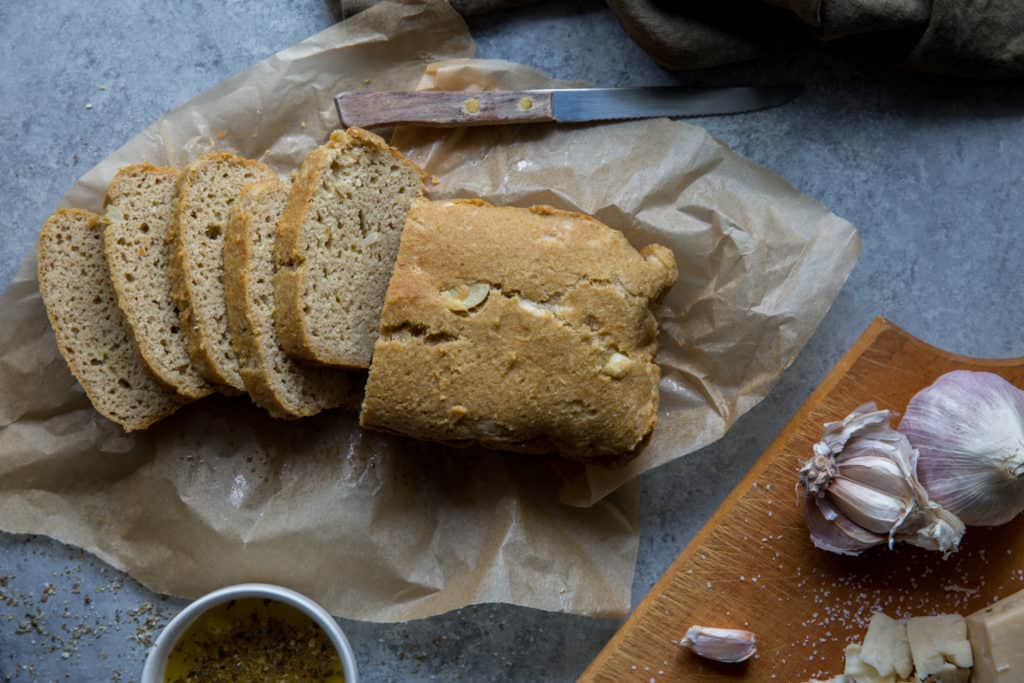keto-garlic-bread-side A half sliced loaf of ket garlic bread on parchment paper with a knife and garlic bulbs and sliced cheese on a wooden cutting board