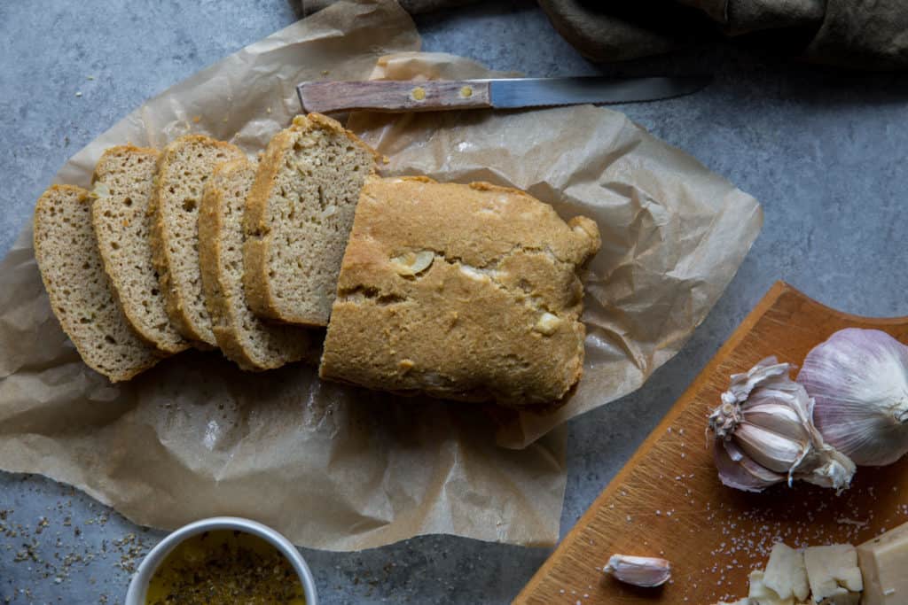 A half sliced loaf of ket garlic bread on parchment paper with a knife and garlic bulbs and sliced cheese on a wooden cutting board