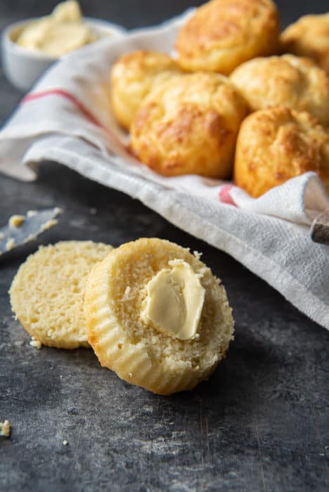 A biscuit sliced up with butter spread on the top half with biscuits and butter in the background on a white and red kitchen towel