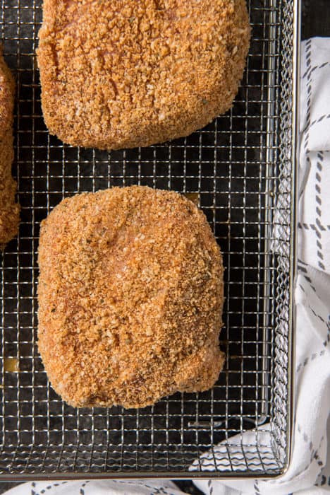 Three pork rind crusted pork chops in an air fryer basket on top of a white and gray kitchen towel