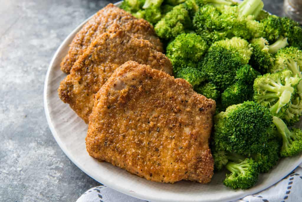 Three air fryer pork chops served with broccoli on a white plate on top of a white and gray kitchen towel