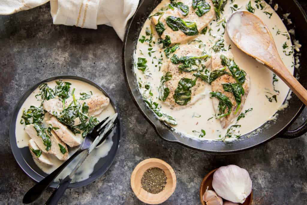 A large cast iron skillet filled with chicken and spinach florentine with a wooden spoon in it and a plate of the chicken and spinach florentine cut up with a knife and fork, a side of black pepper and a garlic bulb