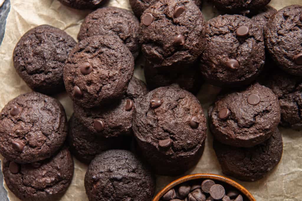 A pile of keto chocolate cookies on brown parchment paper with a bowl of chocolate chips