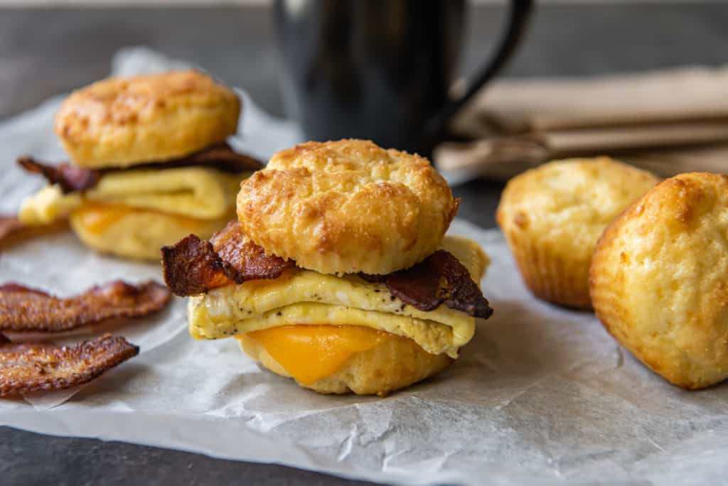 Two Keto breakfast sandwiches that are made with bacon and egg. Extra biscuits and bacon in background