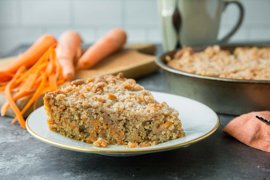 A slice of keto coffee cake on a white plate with shaved carrots, a coffee mug and the rest of the keto coffee cake in the background