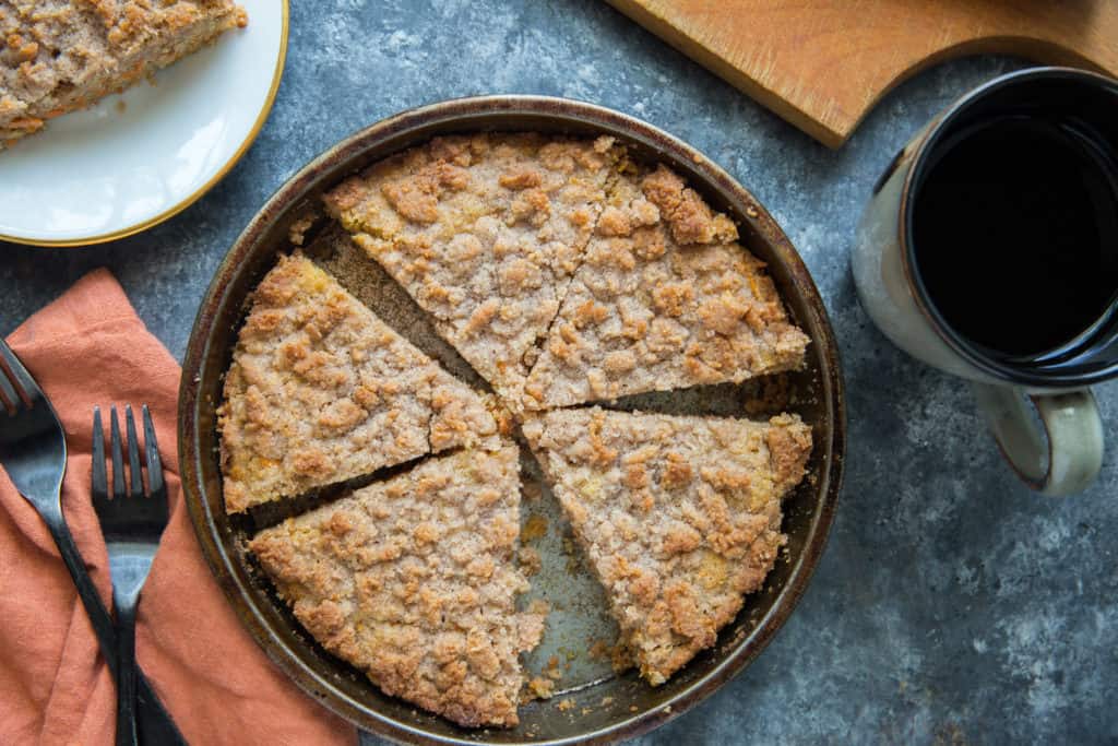 A sliced up keto coffee cake in a cake pan with a coffee mug on the right and forks and a slice of keto coffee cake on a plate on the left