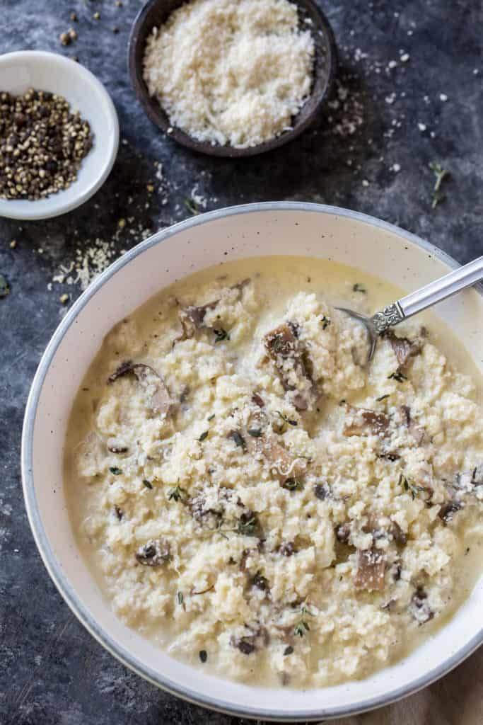 a white bowl of cauliflower risotto with a spoon and a side of black pepper and grate parmesan