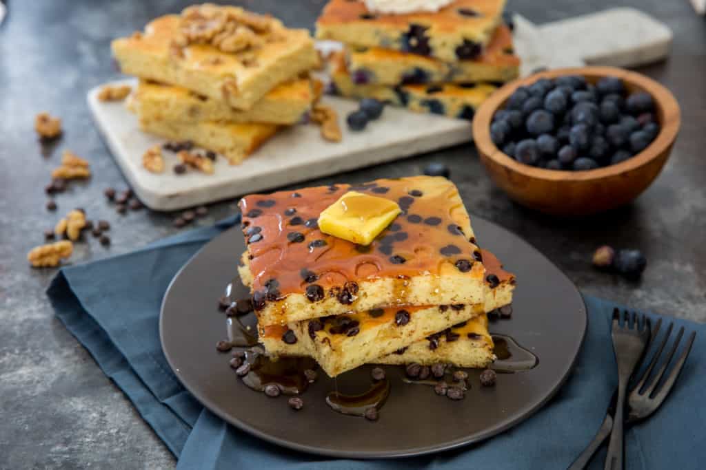 sheet pan pancakes a stack of sheet pan pancakes on a black plate topped with butter and low carb maple syrup with more sheet pan pancakes in the background