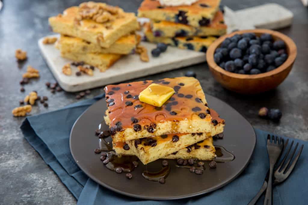 a stack of sheet pan pancakes on a black plate topped with butter and low carb maple syrup with more sheet pan pancakes in the background