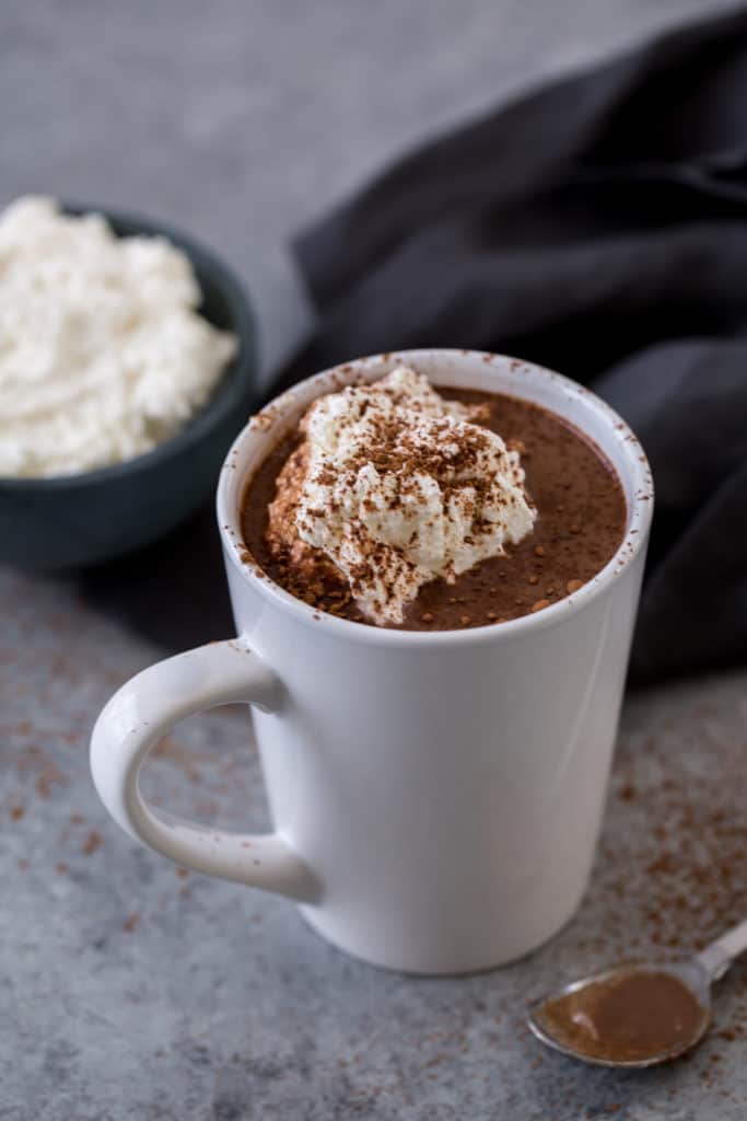 A white mug with cocoa next to whipped cream and a silver spoon