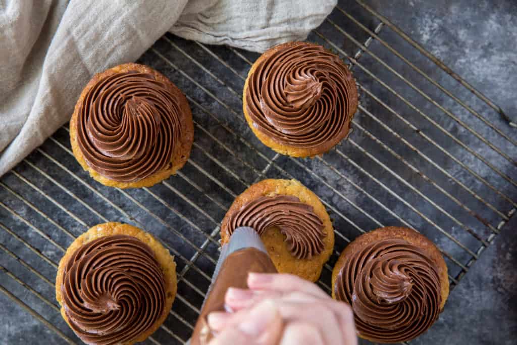 cooked vanilla keto cupcakes being frosted with chocolate frosting on a cooling rack
