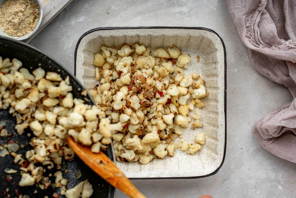 roasted cauliflower being pushed into a baking dish from a skillet with a wooden spoon