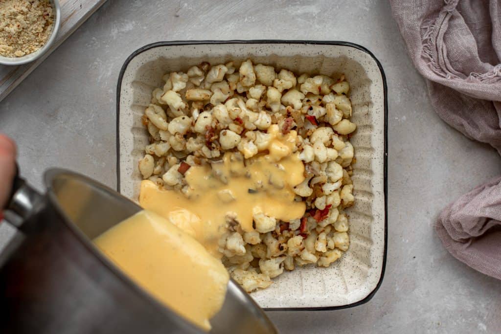 A baking dish filled with cheese sauce and cauliflower next to a lavender colored hand towel. 