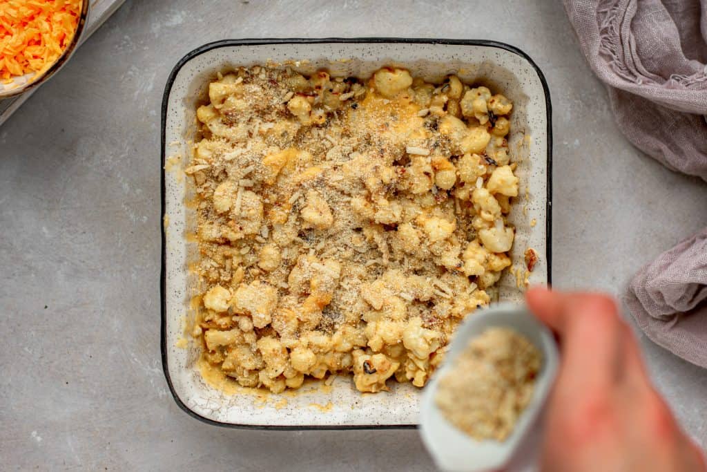 Cauliflower Mac and Cheese Sprinkling crushed pork rinds over top of the mac and cheese next to a bowl of extra cheddar