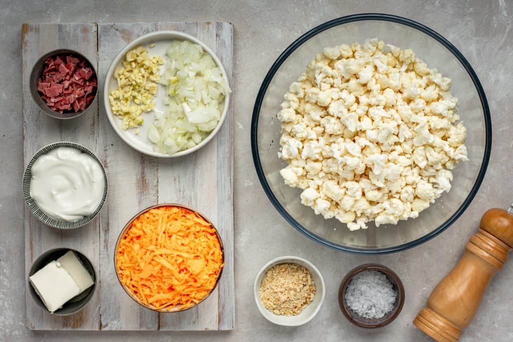 Cauliflower Mac and Cheese All the ingredients for Keto Mac and Cheese in bowls on a grey wood cutting board