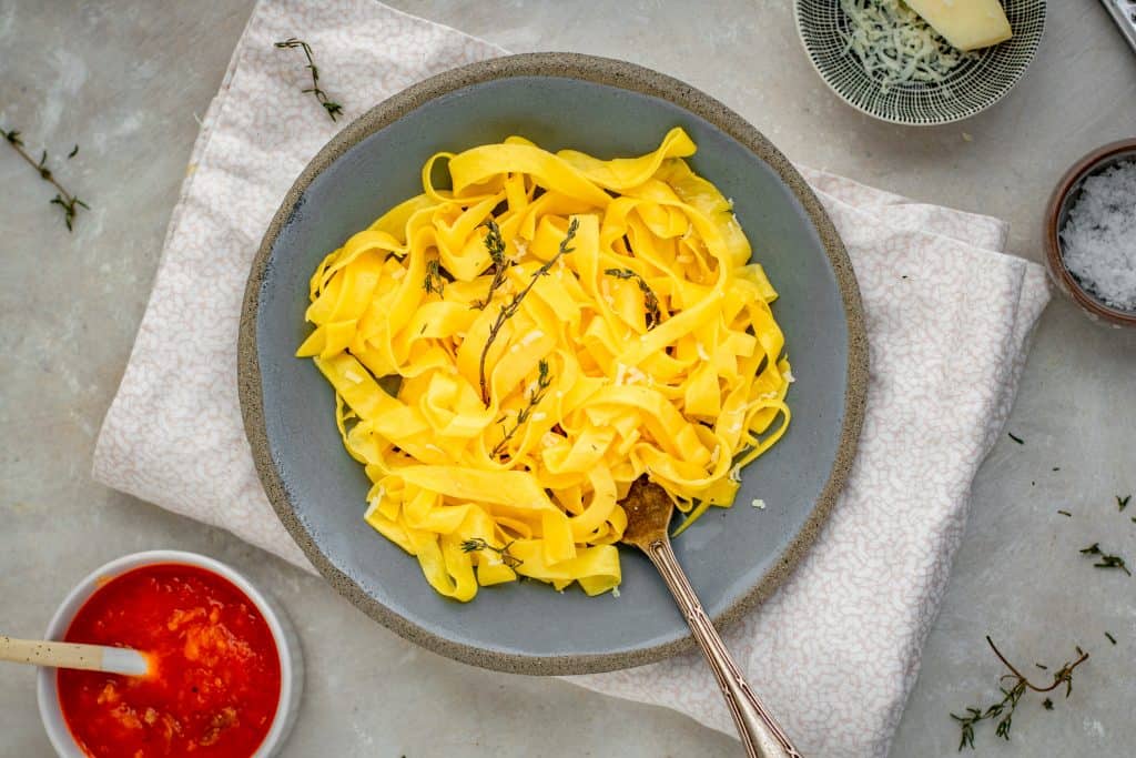 completed pasta with a silver fork next to marinara sauce and grated cheese in ceramic bowls