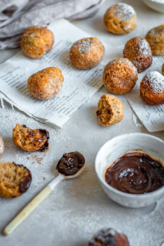 A bowl of chocolate with a spoon dipped in it next to freshly fried donuts.