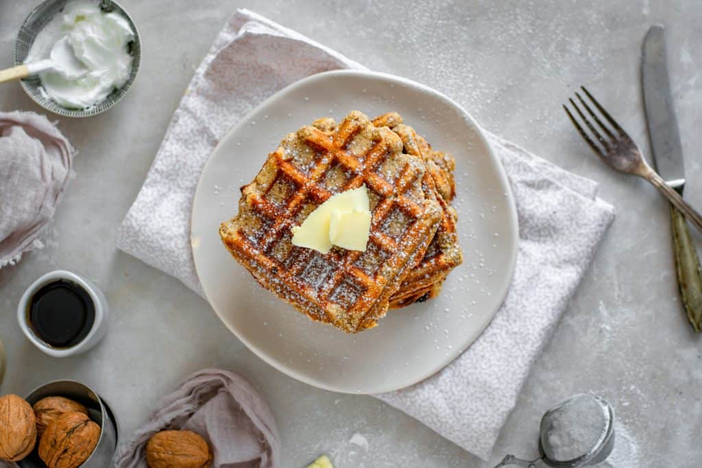 A stack of keto waffles topped with butter next to a container of syrup and whipped cream.