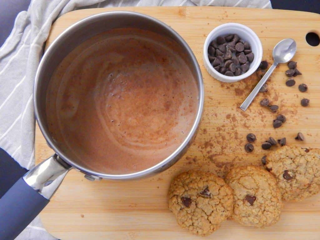 A silver saucepot of hot cocoa next to cookies and chocolate chips. 