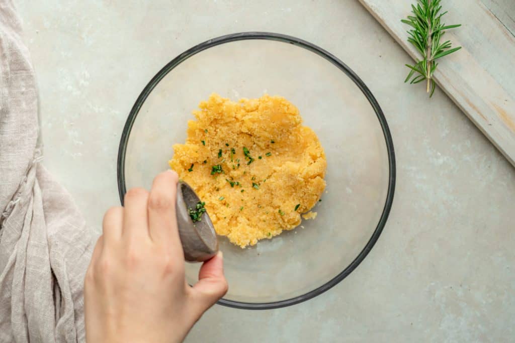 Rosemary being sprinkled into a glass bowl with cheddar cracker dough