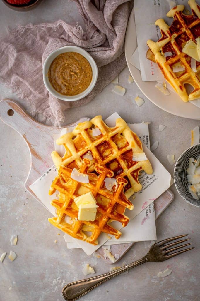 Buttered protein waffles next to coconut flakes, almond butter, and a silver fork. 