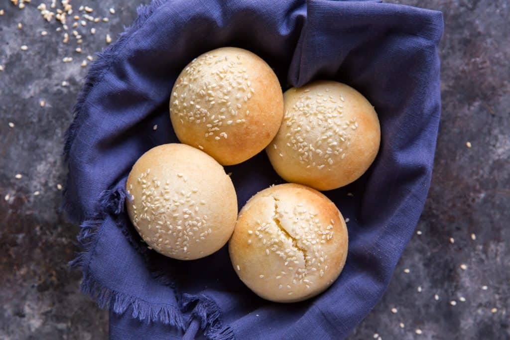 A bread basket lined with a blue cloth.  It's filled with fresh hamburger buns.