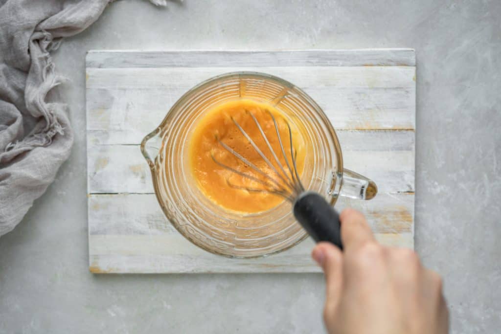 A hand whisking egg yolks in a large glass measuring cup