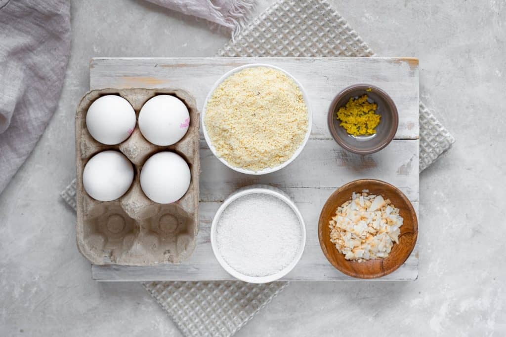 Ingredients for almond cake set out in separate bowls. Eggs, almond flour, erythritol, lemon zest, and sliced almonds