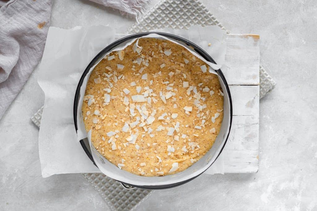 Overhead view of whole almond flour cake topped with coconut flakes in a cake pan. It is sitting on a rustic white board and a piece of parchment paper.