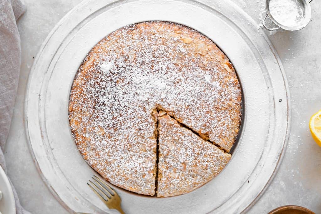 A whole almond cake on a white serving tray with a piece cut out. Lemon and powdered sugar duster next to it.