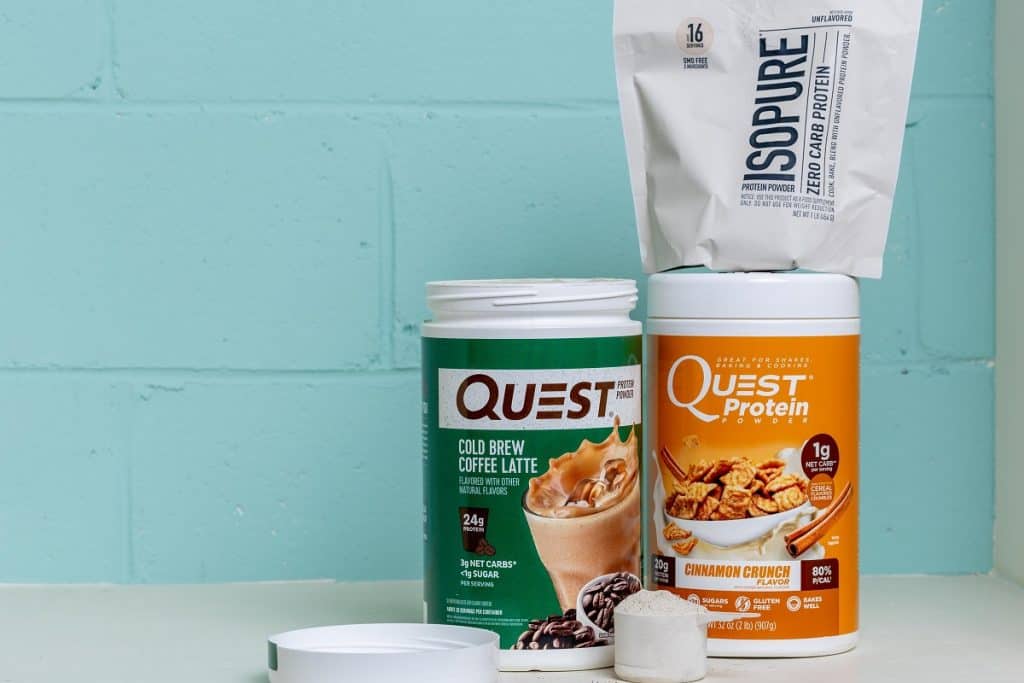 Two canisters of quest and one of isopure protein powders with a scoop of protein sitting in front of them.