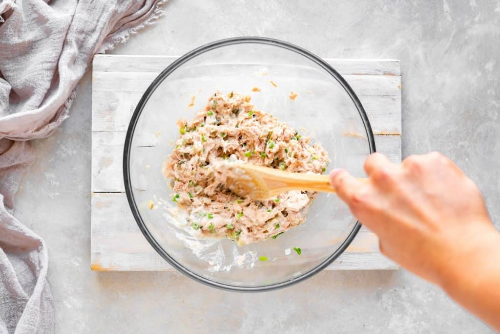 overhead shot of a wooden spoon mixing all of the ingredients for tuna salad together in a glass bowl
