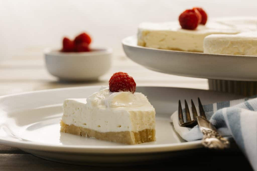 Slices of cheesecake with a raspberry on top with more raspberries and the rest of cake in background