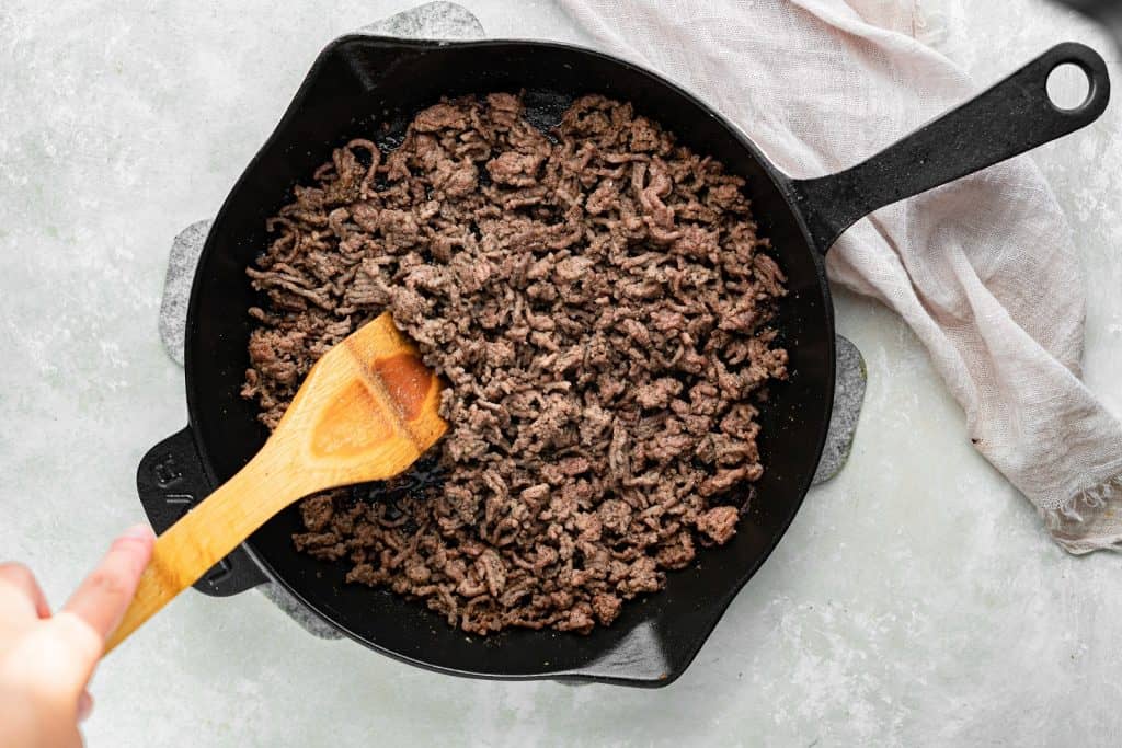 Ground beef in a cast iron skillet, being mixed with a wooden spoon.