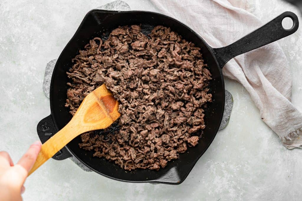 big mac salad Ground beef in a cast iron skillet, being mixed with a wooden spoon.