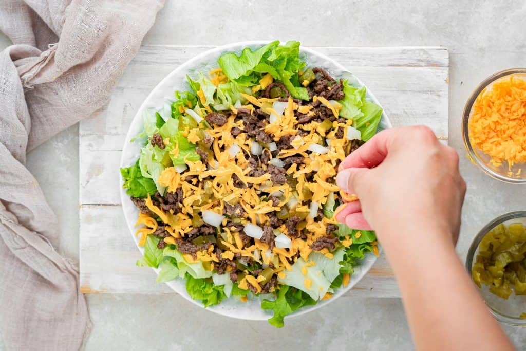 Sprinkling cheese on a Big Mac salad. Salad is in a white bowl sitting on a rustic white washed board.