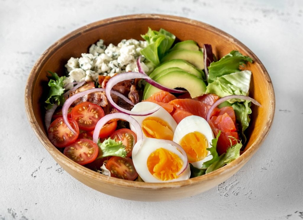A wooden bowl with mixed greens, salmon, tomato, eggs, and bacon sitting on a concrete tabletop